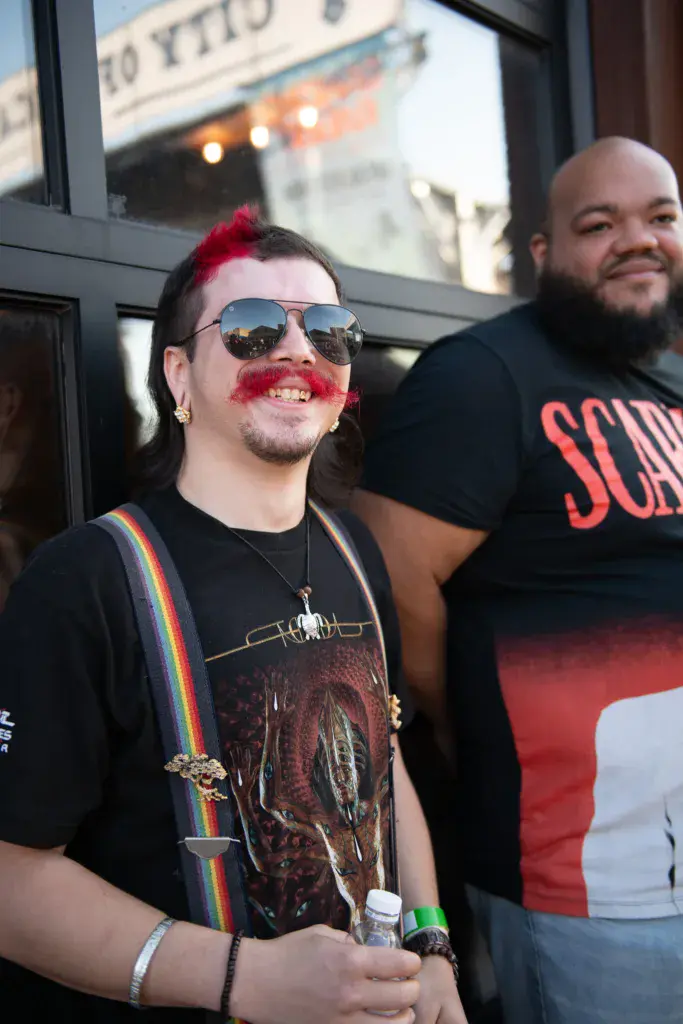 Two festival attendees smile at the camera in front of a venue storefront, one wearing sunglasses and red face paint.