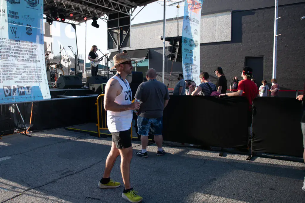 Festivalgoers stand near a stage barrier while a performer plays guitar on stage at an outdoor music venue.