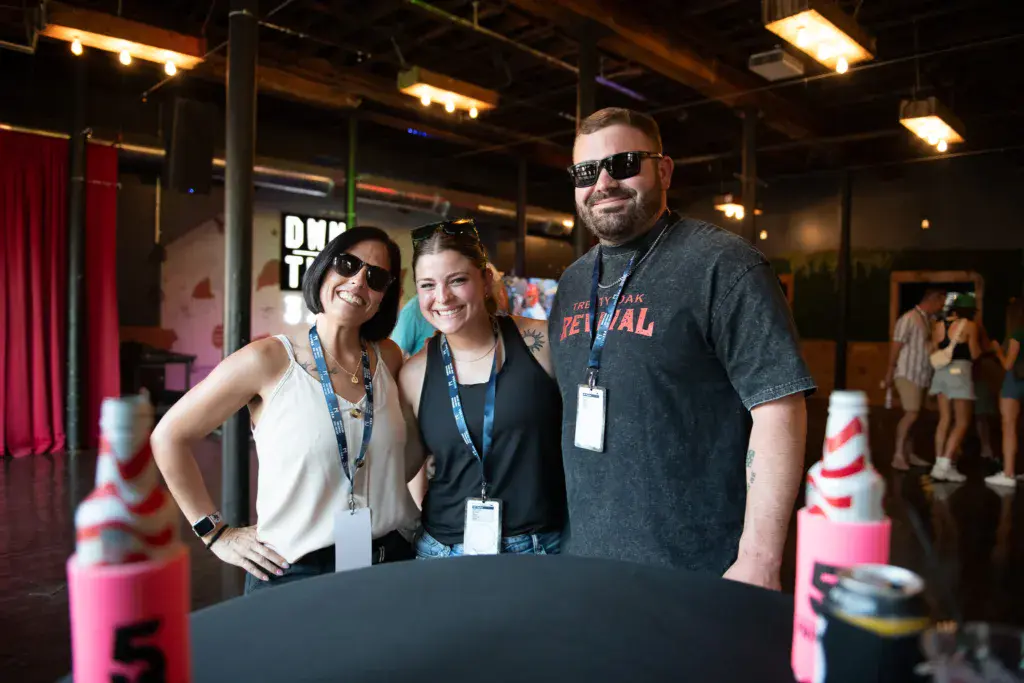 Three festival staff members wearing credential badges smile together at an indoor venue booth.