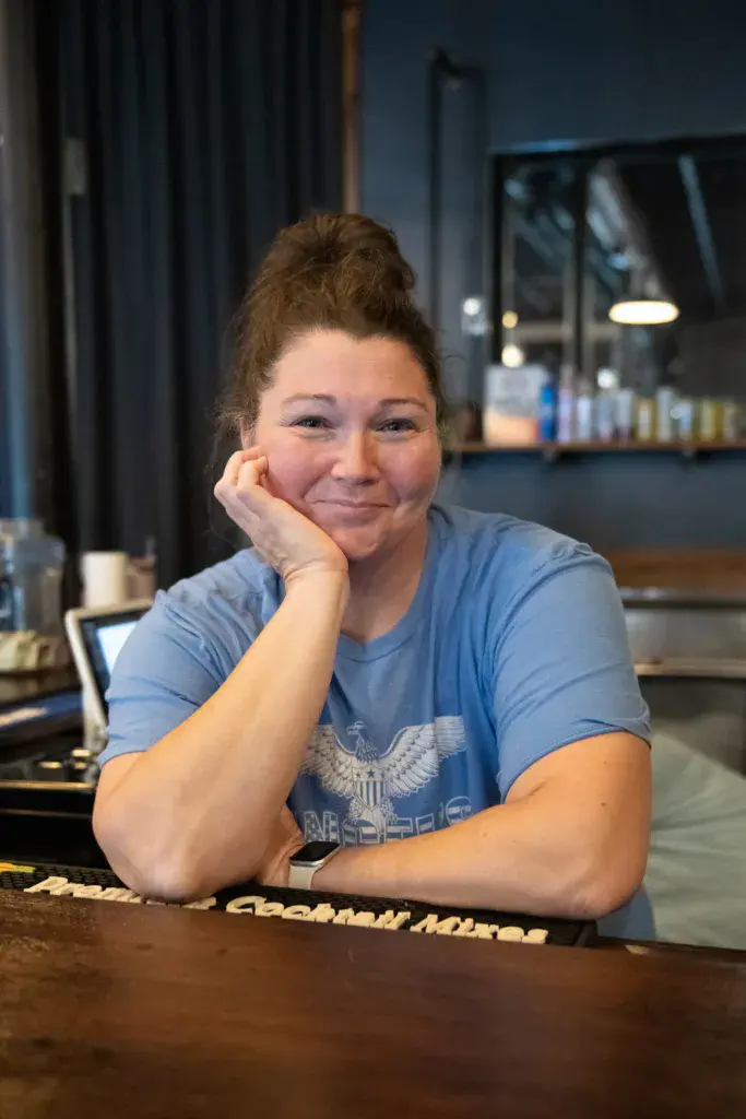 A person in a blue graphic t-shirt rests their chin on their hand while leaning against a bar counter.