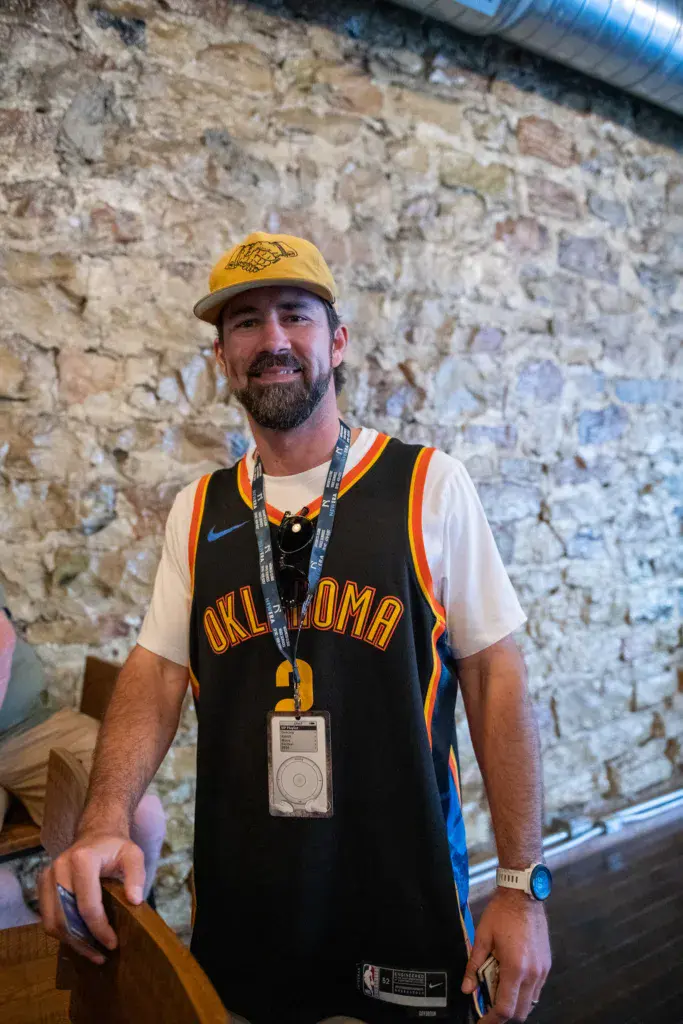 Person wearing a yellow cap and Oklahoma basketball jersey with festival credential badge stands against a rustic stone wall.