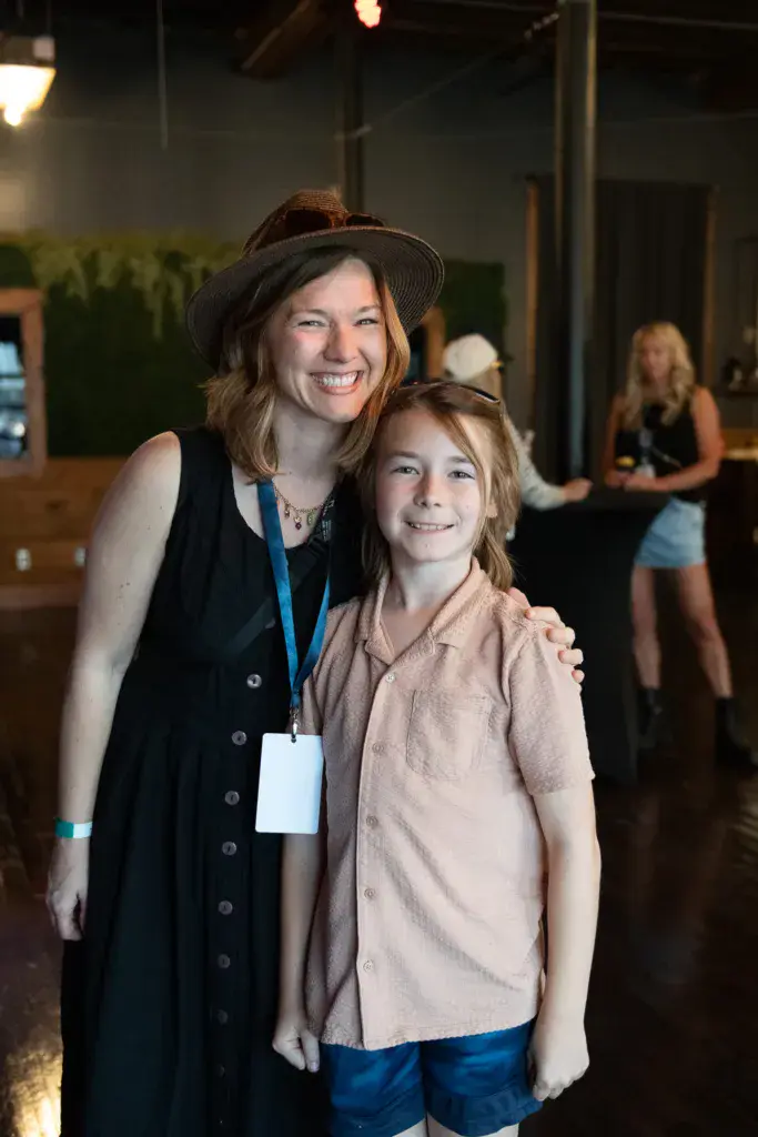 Two people smiling at the camera, one wearing a black hat and lanyard at an indoor festival venue.