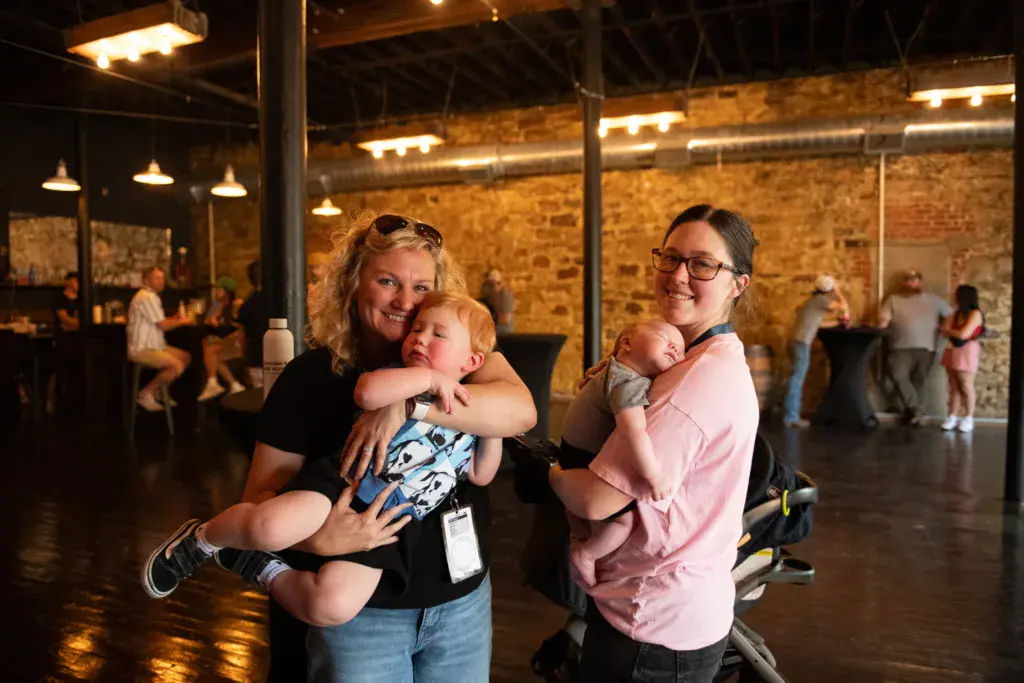 Two adults holding young children smile at the camera inside a brick venue at night.