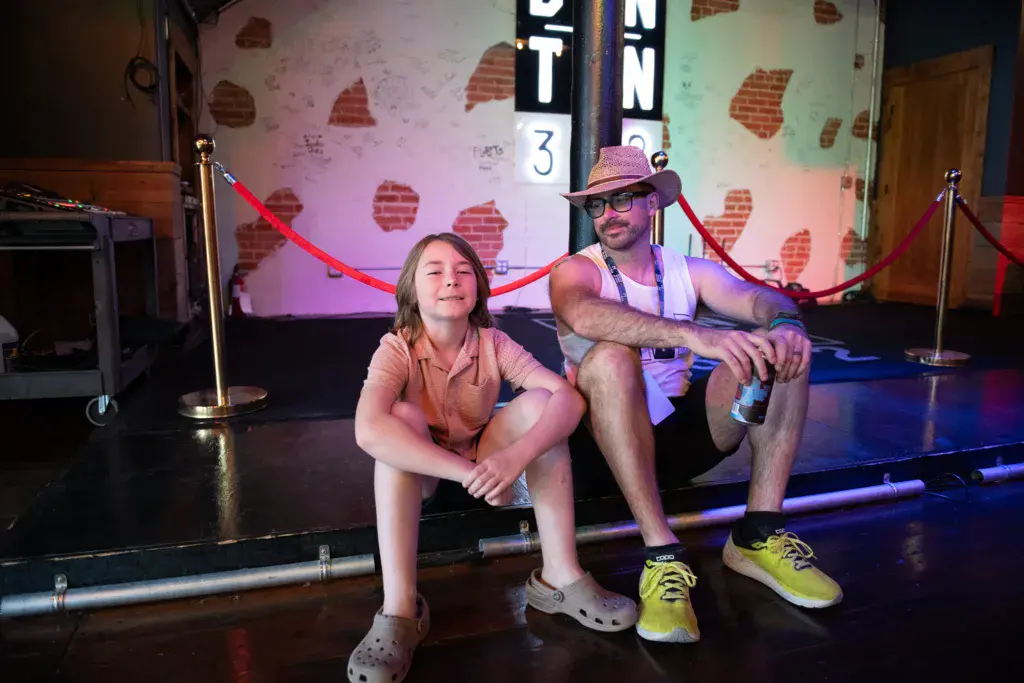 Two people sitting on the stage floor in front of red velvet ropes and illuminated signage at an indoor venue.
