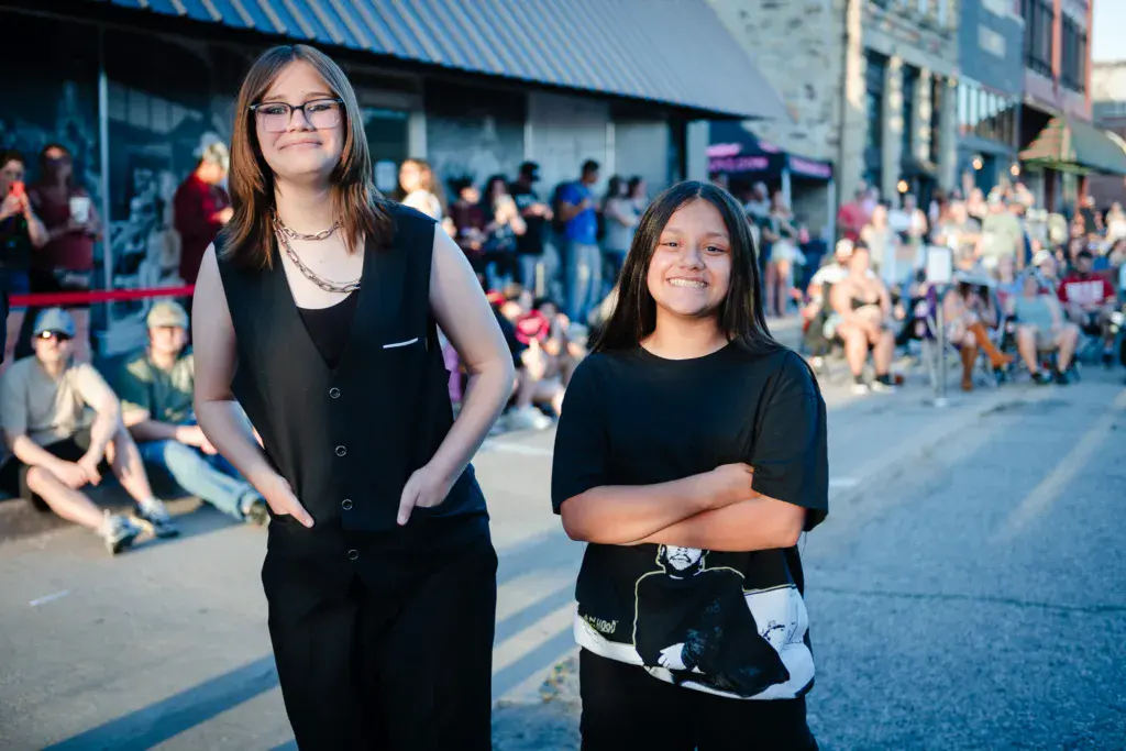Two young women in black clothing stand smiling in front of a crowded outdoor festival venue.