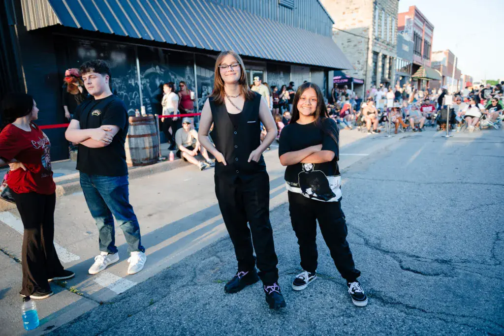 Young people in black clothing stand on a street during an outdoor festival with crowds and buildings in the background.