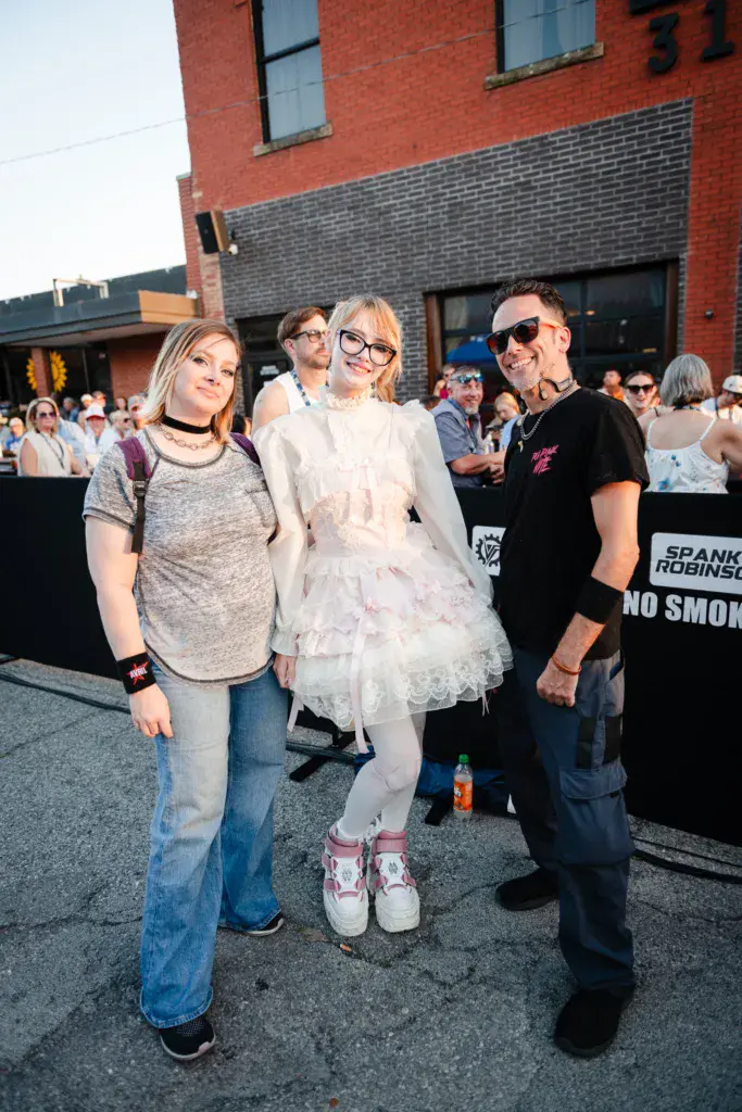 Three festival attendees pose together in a crowded outdoor venue in front of a brick building.