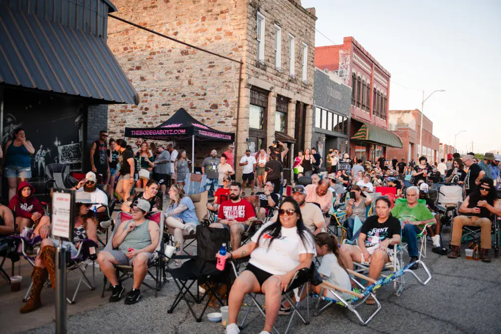 A large crowd of festival-goers seated in lawn chairs fills a downtown street lined with historic brick buildings.