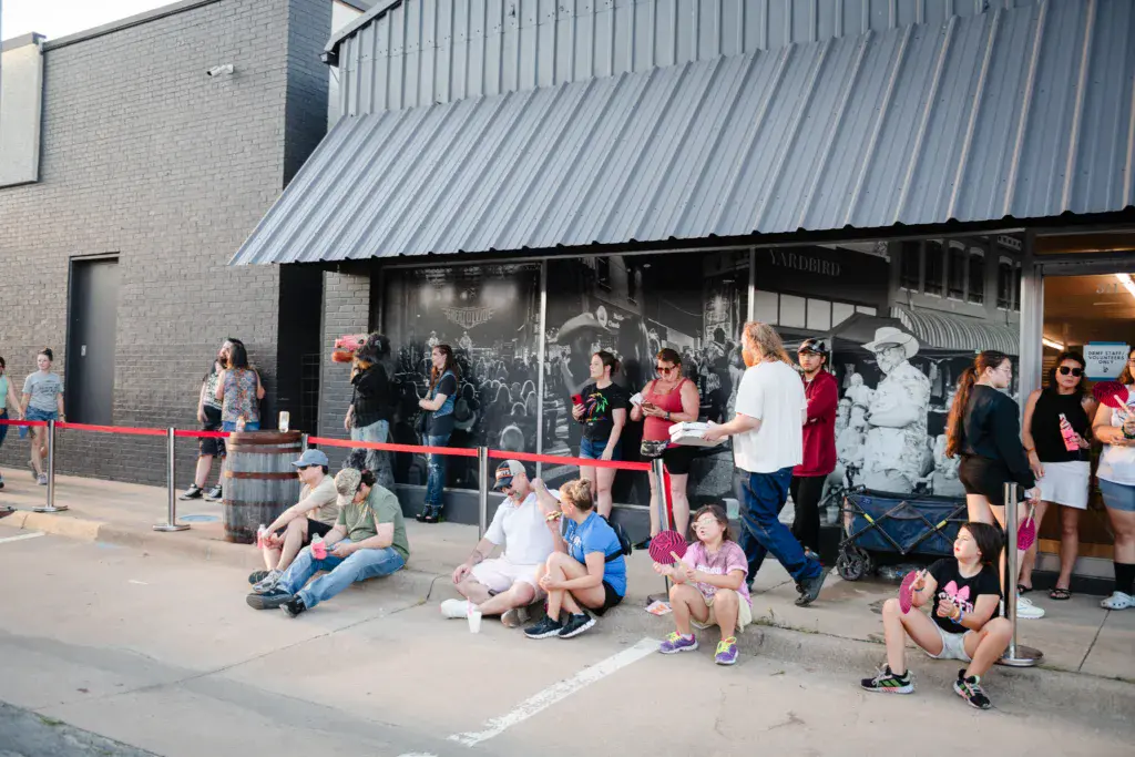 Festival attendees wait in line outside a venue building with corrugated metal roofing.