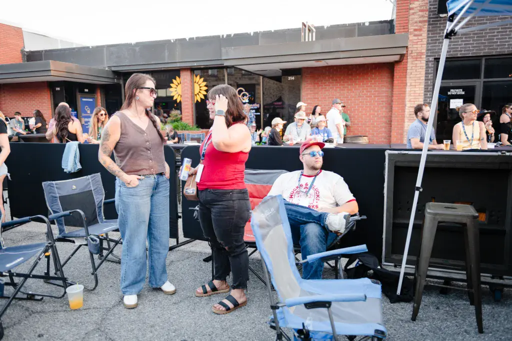 Festival attendees socialize in an outdoor venue area with brick buildings and food service in the background.