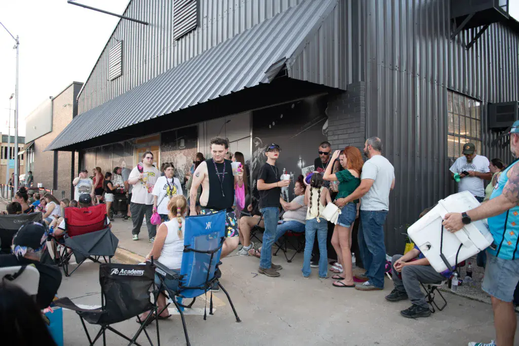 Festival attendees gather outside a large metal venue building, some seated in lawn chairs while others stand and mingle.