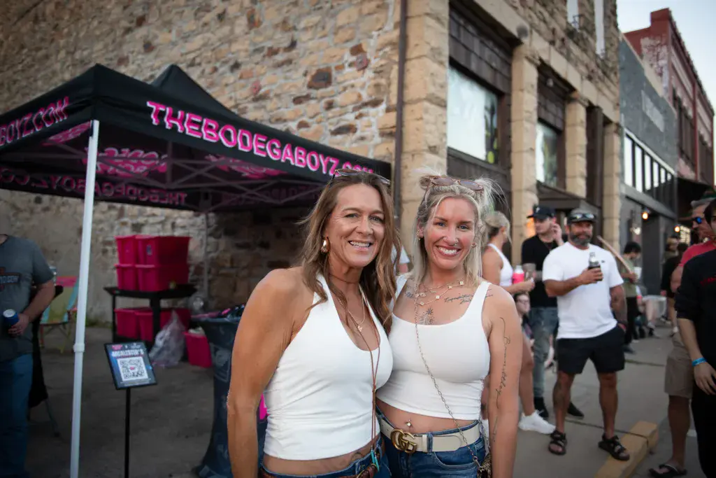 Two attendees smiling at the camera in front of a sponsor tent at an outdoor music festival venue.