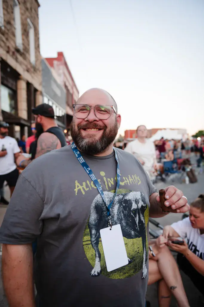 A festival attendee wearing glasses and a band t-shirt smiles at the camera while wearing a festival credential.