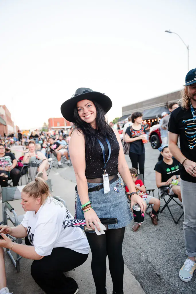Festival attendees enjoying the outdoor music event, with one person wearing a black hat smiling at the camera while surrounded by seated crowd members.