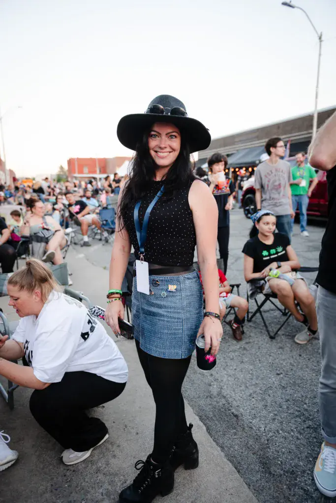 A smiling person wearing a black wide-brimmed hat and lanyard stands among festival-goers in a crowded outdoor venue.