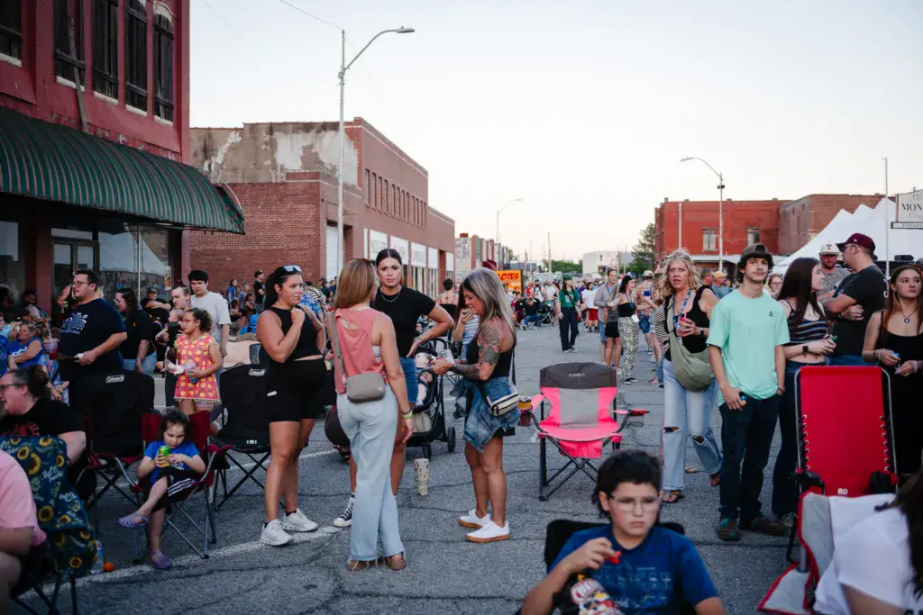 A large crowd of festival attendees gathers in a downtown street lined with brick buildings during daytime.