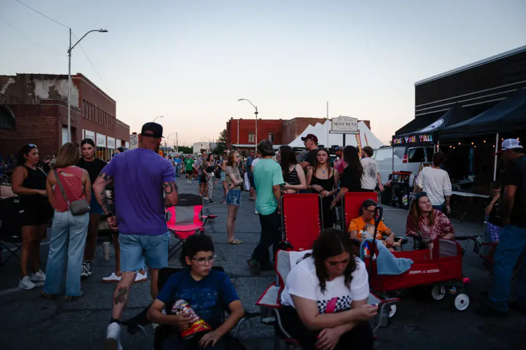 A diverse crowd gathers on a street at dusk near food vendors and historic brick buildings during an outdoor music festival.