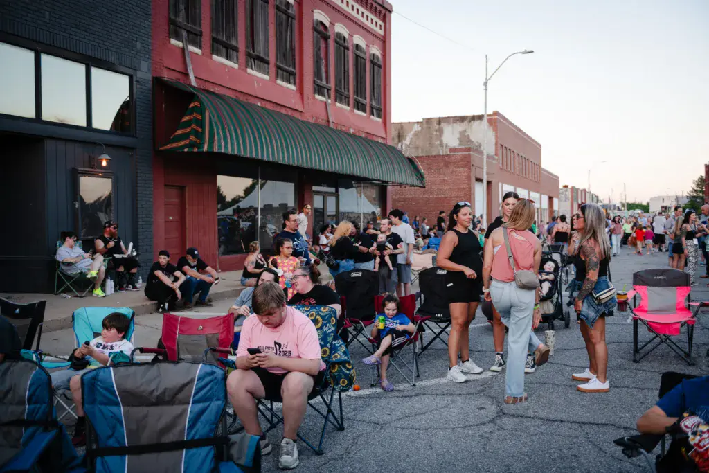 Crowd of festival attendees seated and standing in a street lined with brick buildings at evening time.