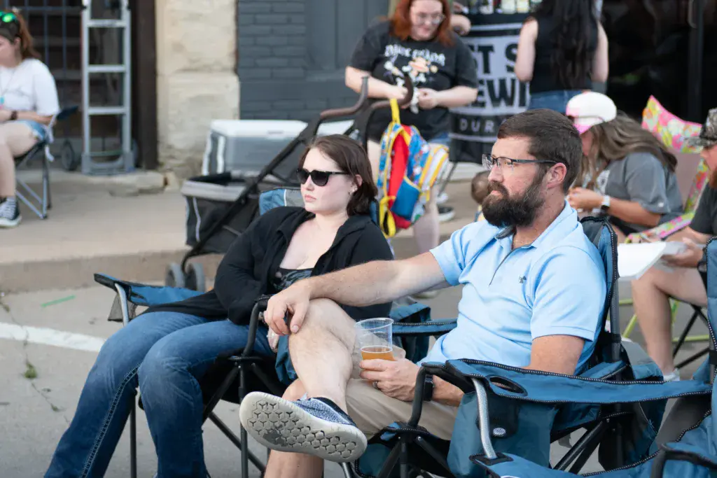 Festival attendees sitting in folding chairs watching the stage, with colorful decorations and other spectators visible in the background.