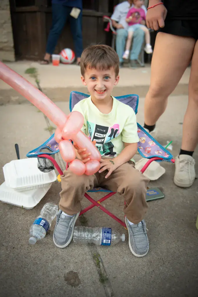 Young child sitting in a folding chair holding a pink balloon animal at an outdoor festival.