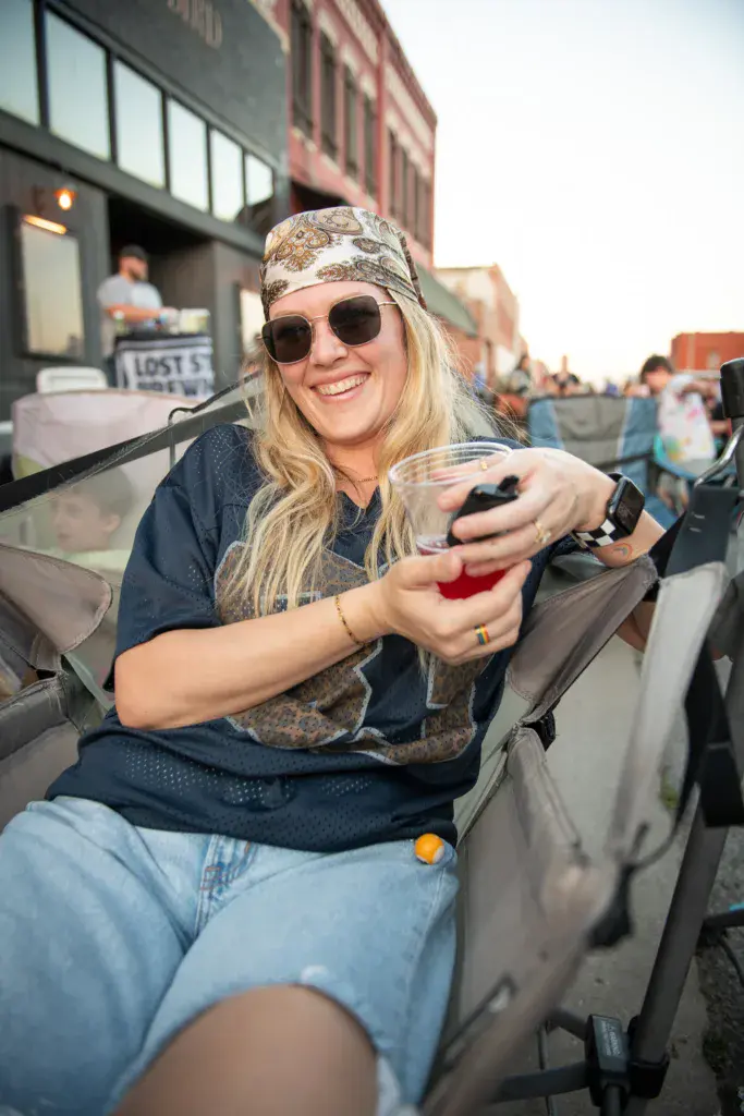 A festivalgoer wearing sunglasses and a bandana holds a red beverage while smiling at an outdoor music event.
