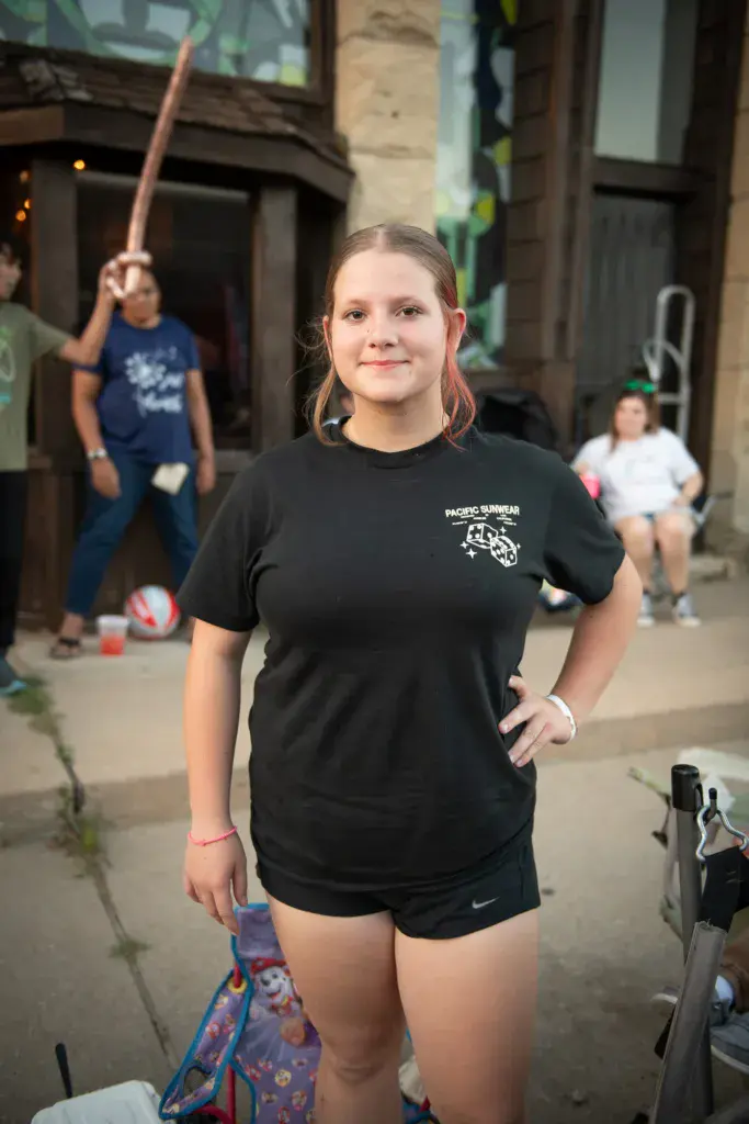 A young person in a black t-shirt smiles at the camera while festival-goers play in the background.