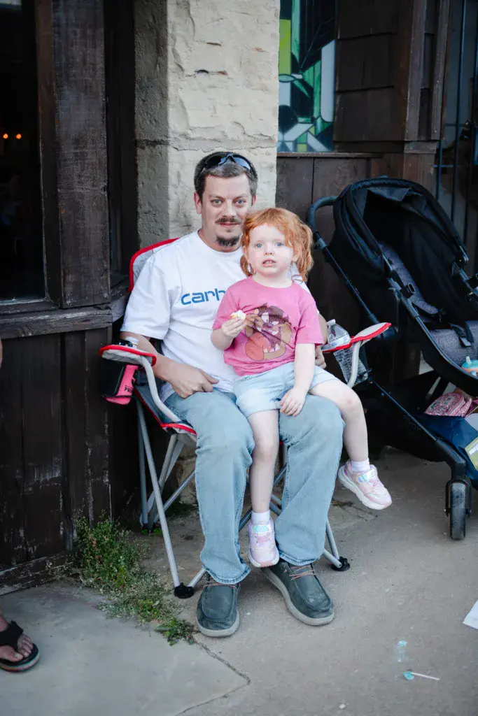 An adult and young child sit together on a camping chair at an outdoor venue, taking a break during the festival.