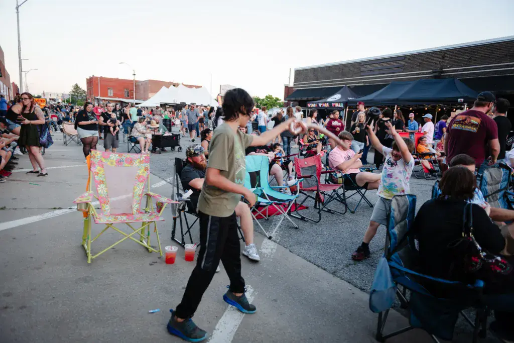 Festival attendees gather in a parking lot with camping chairs, one person dancing in the foreground.
