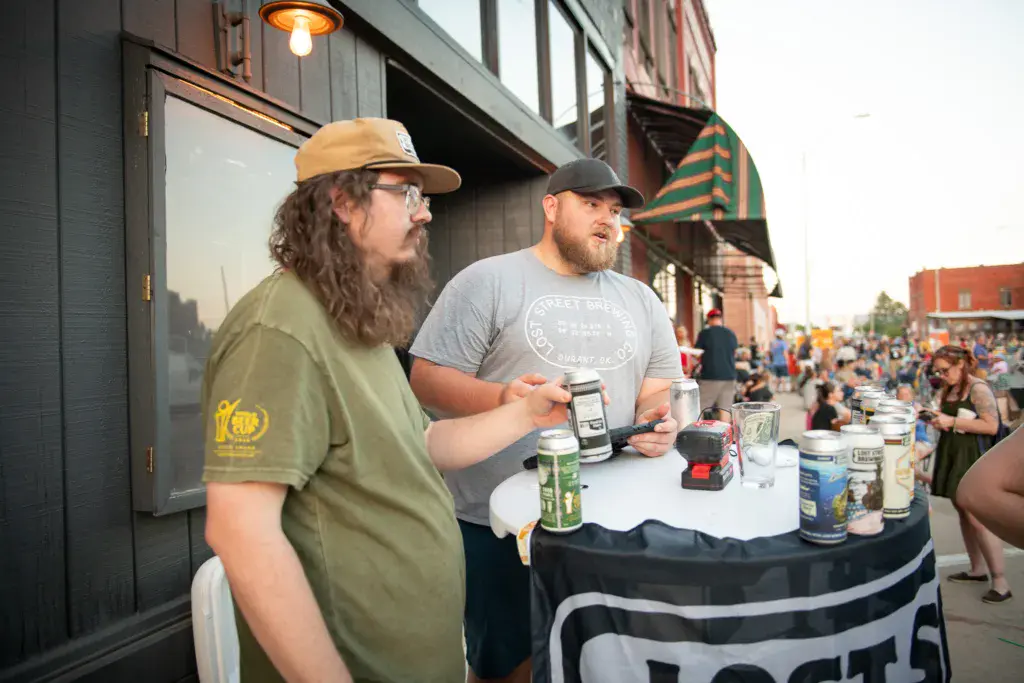 Two people at a festival table with beverages and merchandise, with crowds and buildings visible behind them.