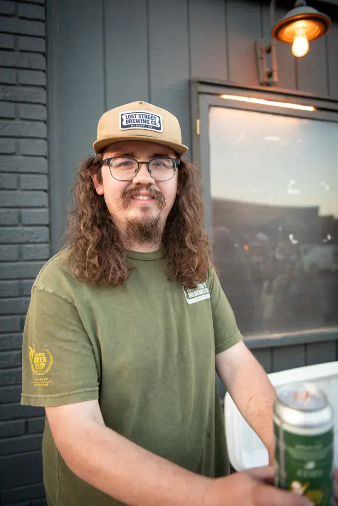 Person wearing Lost Street Brewing Co. cap and olive green t-shirt smiles at camera in front of industrial venue interior.
