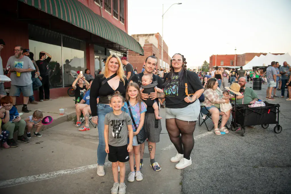 A group of adults and children smile for the camera at an outdoor festival in front of brick buildings.