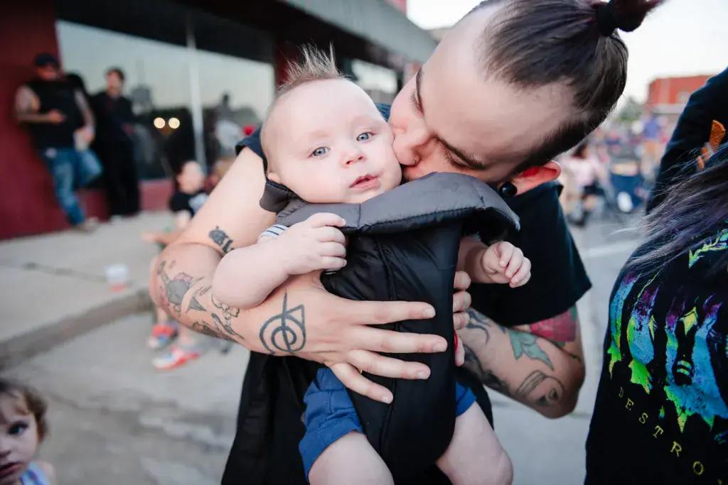 An adult with tattooed arms holds and kisses a smiling infant at the Dancing Rabbit Music Festival.