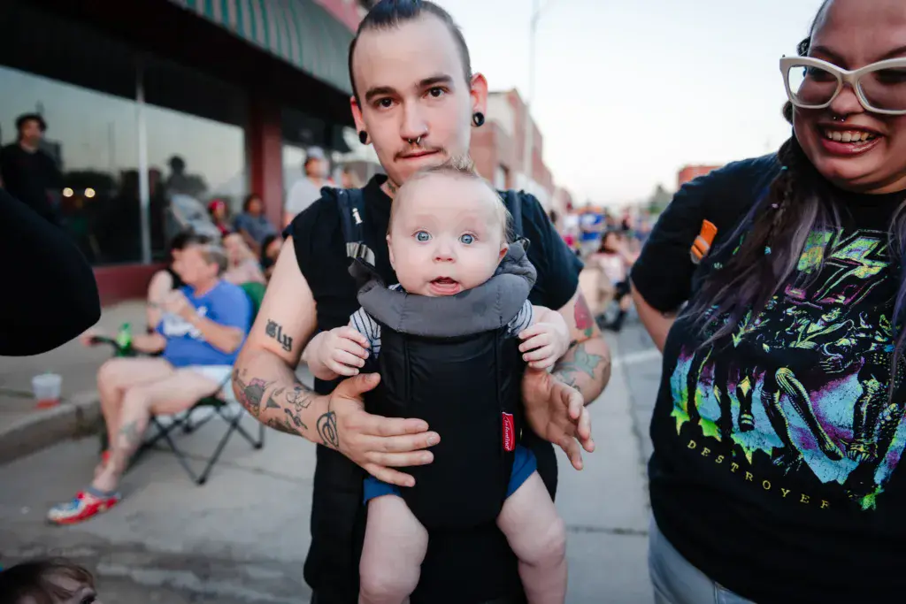 A parent holding a smiling infant at an outdoor music festival surrounded by festivalgoers.