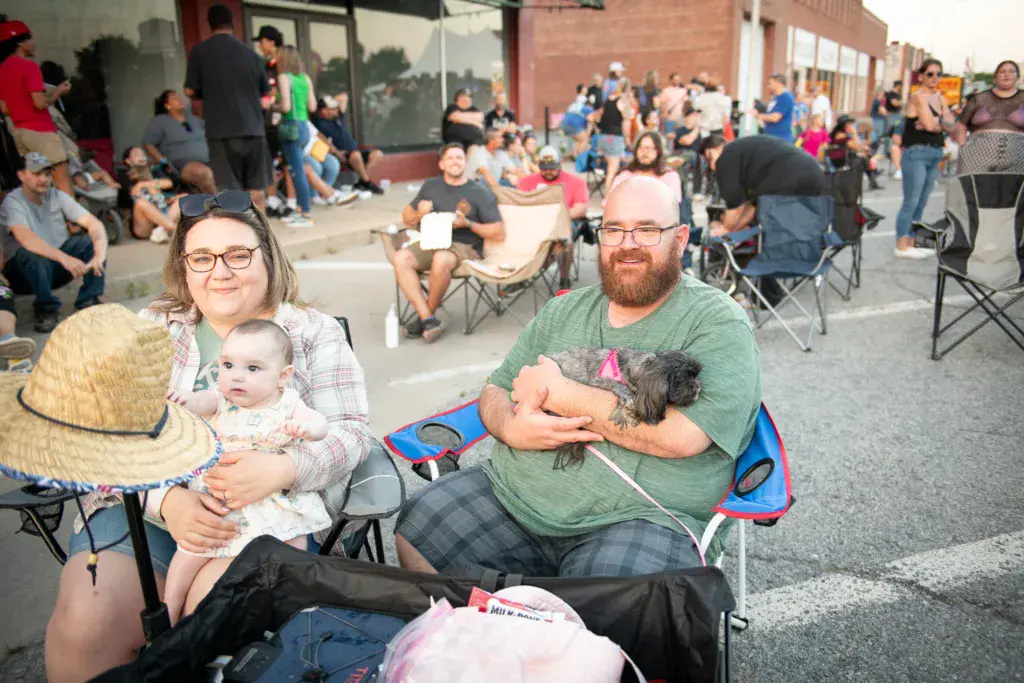 Festival attendees sit in lawn chairs in an outdoor venue, with a woman holding an infant and a man holding a small dog in the foreground.