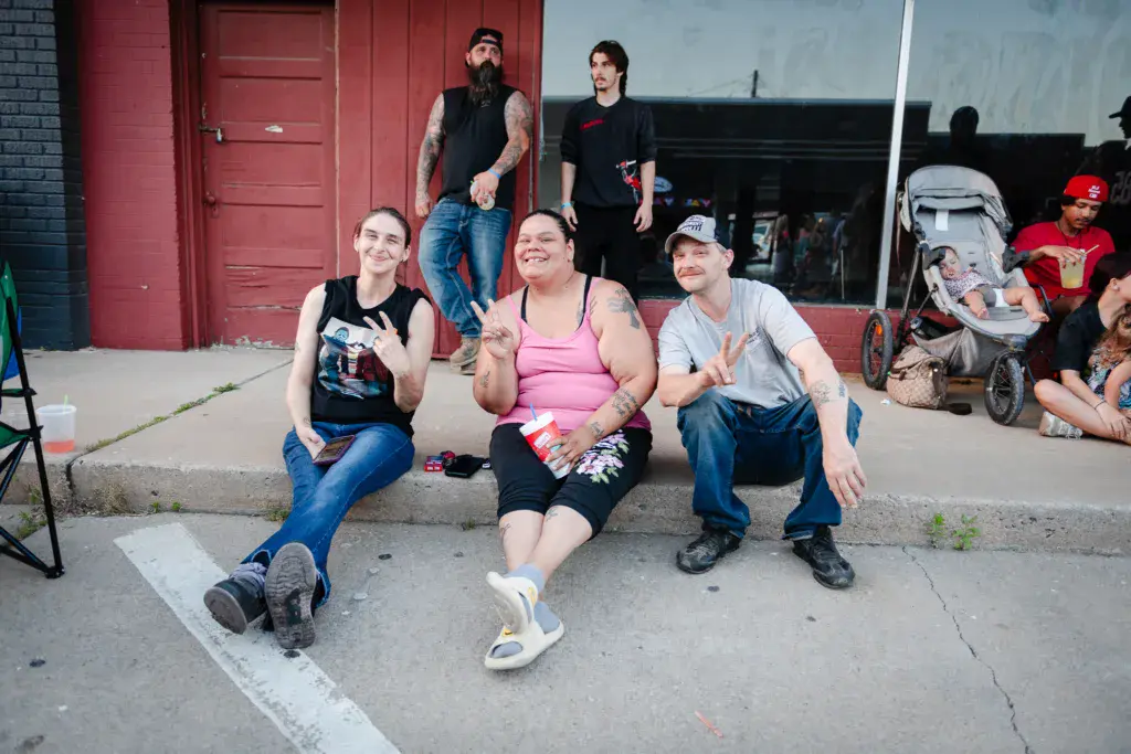 Festival-goers sitting on a curb at an outdoor music event, smiling and enjoying snacks.