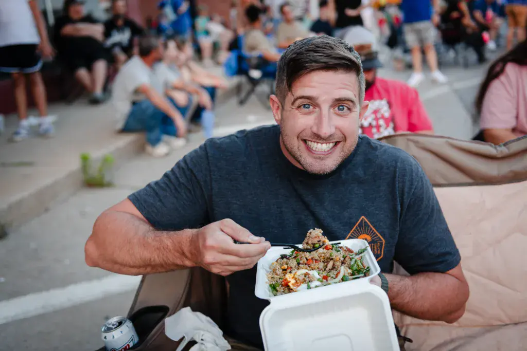 A smiling person holding a takeout container of food at an outdoor festival venue with crowds and camping in the background.