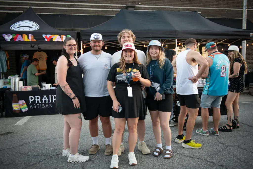 Group of festival attendees posing together in front of a vendor tent at an outdoor music festival.