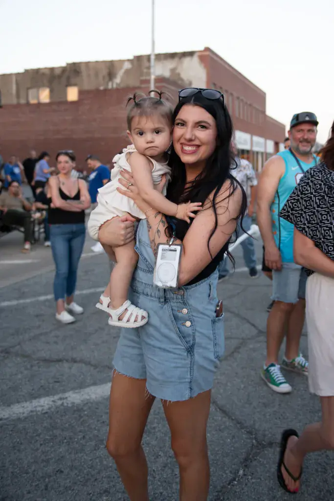 A woman holding a young child stands in a festival crowd near a brick building, smiling at the camera.