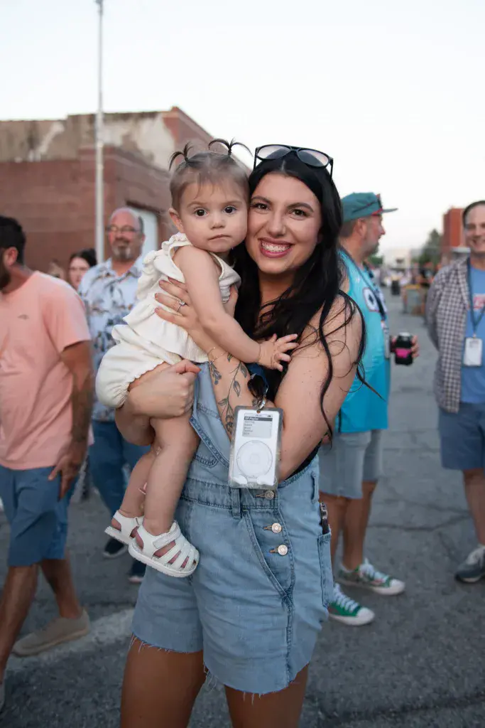 A woman holding a young child smiles at the camera while wearing a festival wristband and sunglasses, surrounded by festival-goers.
