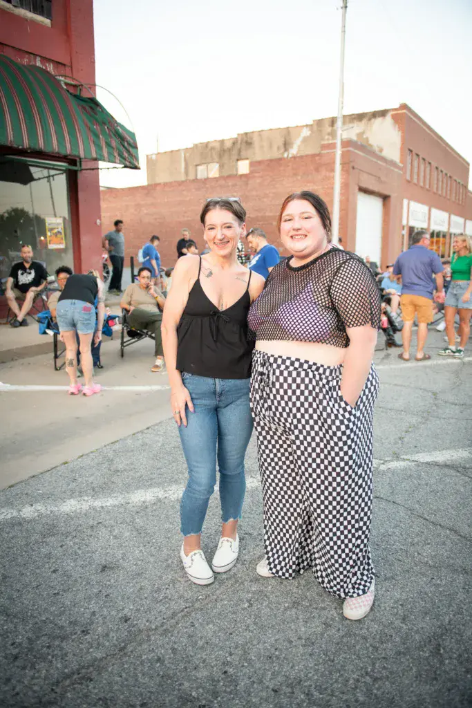 Two smiling festival attendees pose together in front of a brick venue building with crowds behind them.