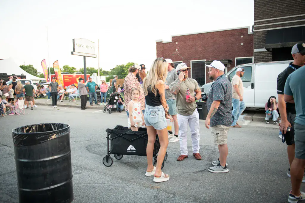 Festival attendees walk through a parking lot venue area with food vendors and signage visible in the background.