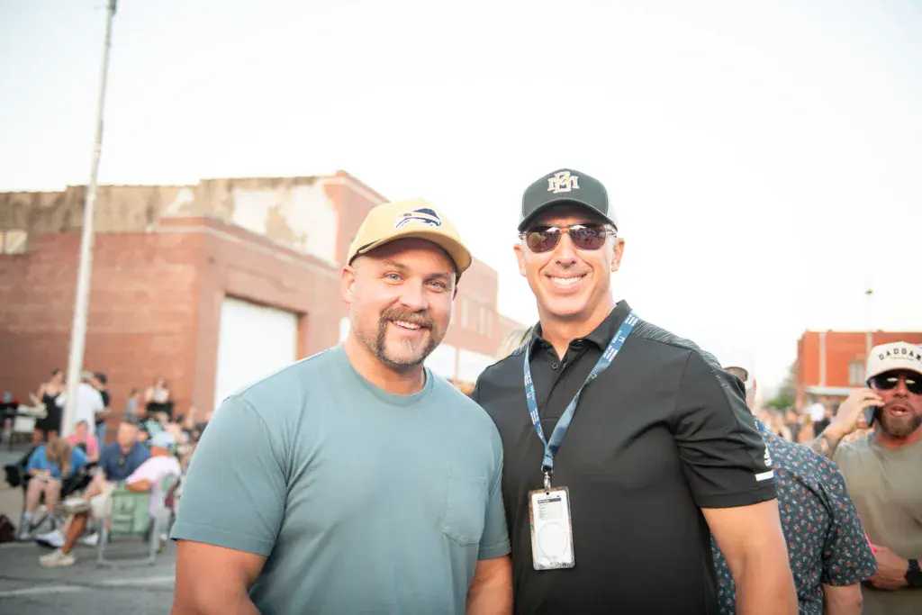 Two men smile at the camera at an outdoor festival venue with brick buildings and crowds visible in the background.