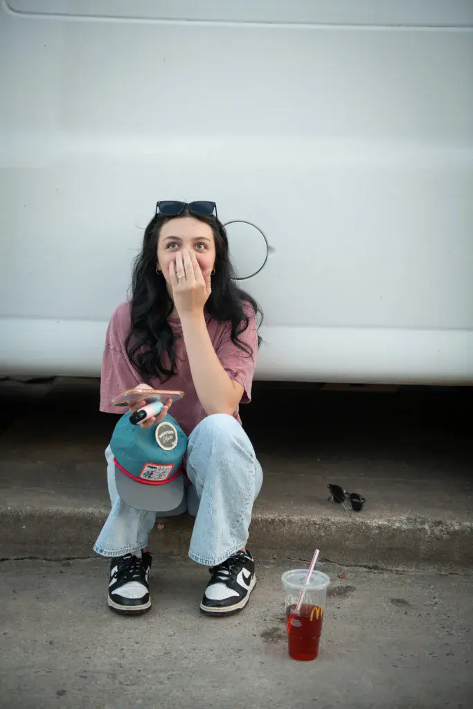 A person in a mauve shirt and jeans sits crouching against a white vehicle, holding a colorful baseball cap and a beverage.