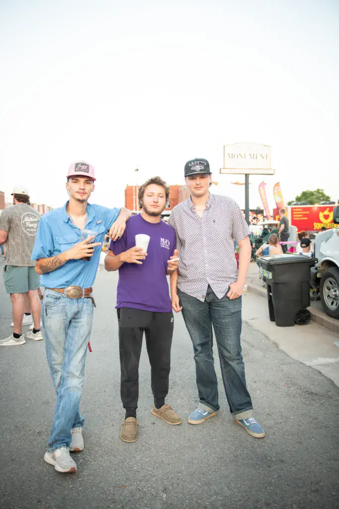 Three men stand together at an outdoor festival with venue signage and crowd visible in the background.