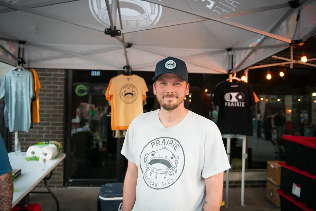 A person wearing a Prairie brewery t-shirt and cap stands under a vendor tent at an outdoor festival.