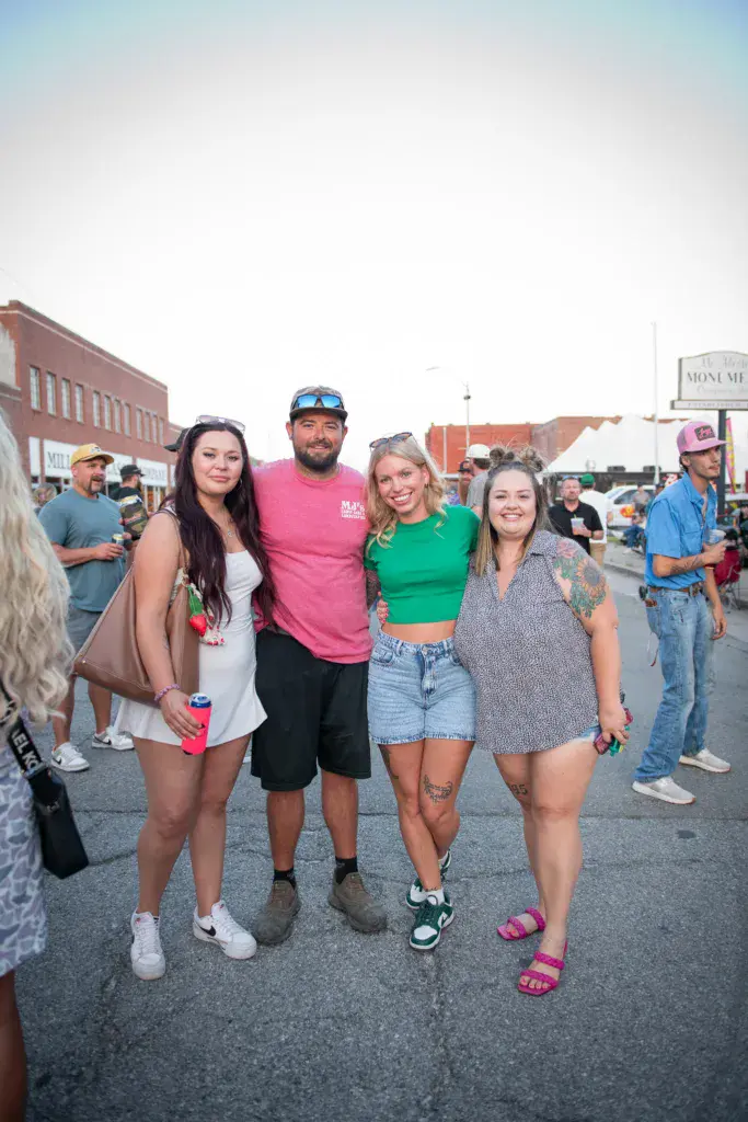 A group of five people pose together at an outdoor festival with historic brick buildings and vendor booths in the background.