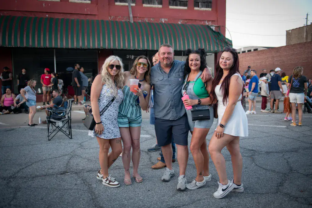 A group of five people smile for a photo in an outdoor festival courtyard with crowds and brick buildings in the background.