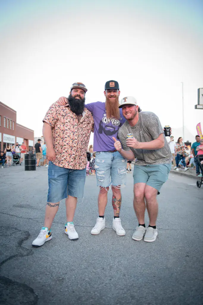 Three attendees pose together on the festival grounds wearing casual summer attire and caps.