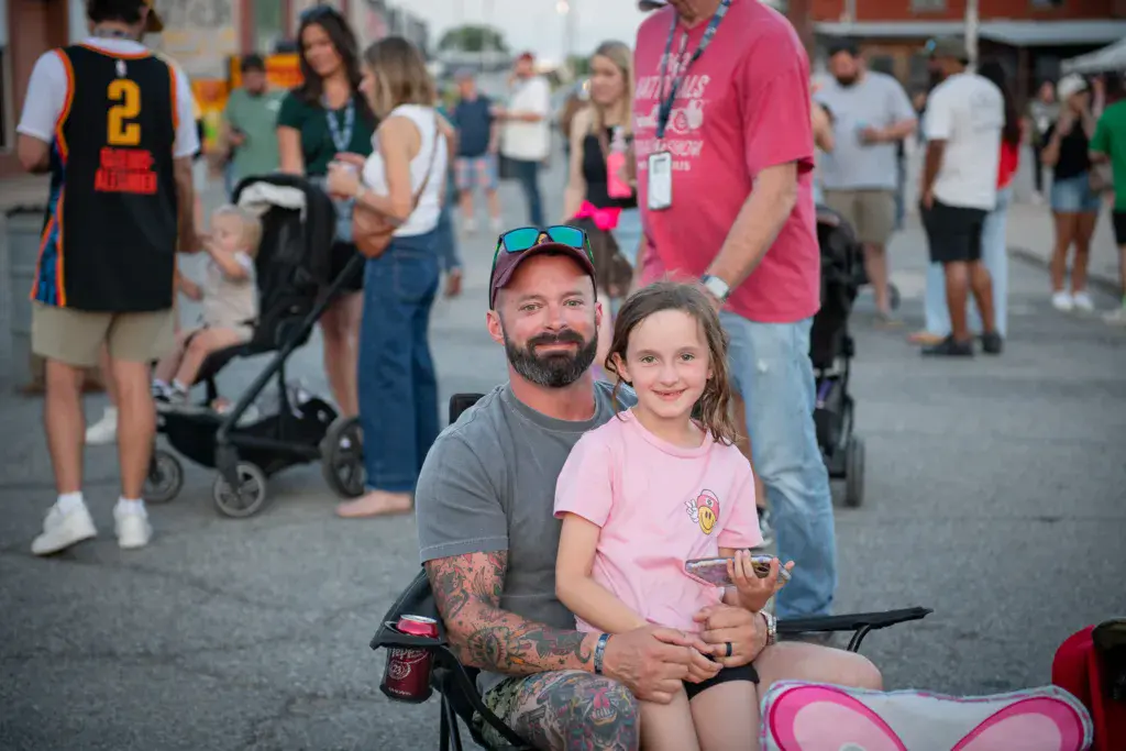 A smiling adult and child sit together in chairs at an outdoor festival surrounded by a crowd of festival attendees.