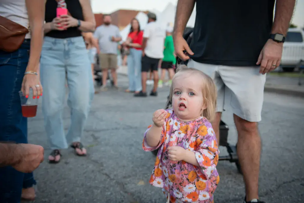 A young child in a colorful floral outfit stands among festival attendees on a paved area.